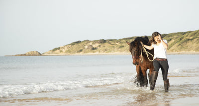 Pferd am Strand mit Reiterin, die mit wasserfesten Reitstiefeln durchs Wasser läuft